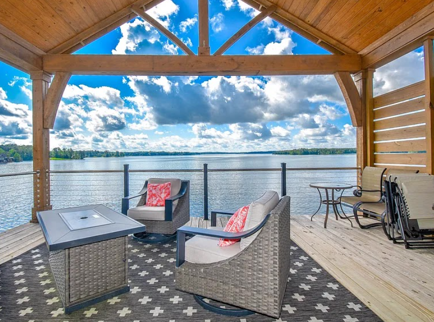 Covered lakeside patio with wicker seating, patterned rug, and a view of calm water and blue sky