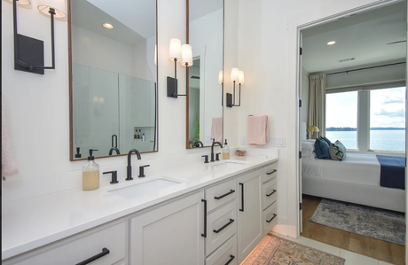 Bright bathroom vanity with double sinks, mirrors, wall sconces, and a doorway to a bedroom with ocean view