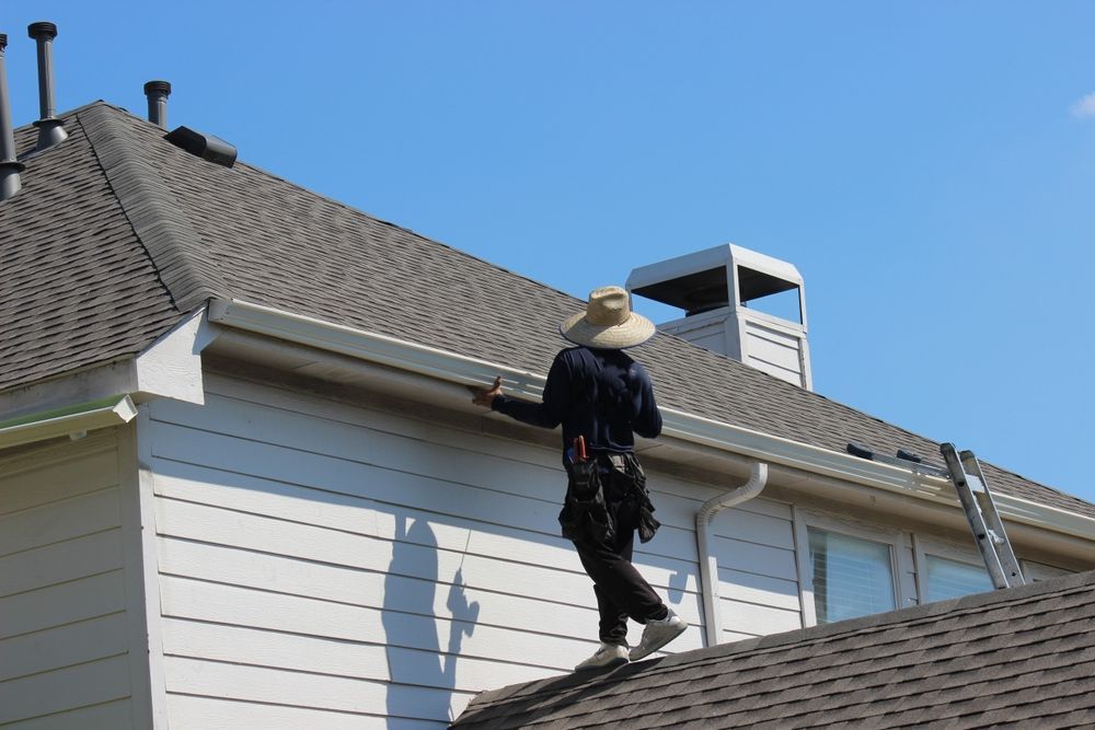 Worker on a house roof inspecting or repairing gray shingles beside a white house wall.