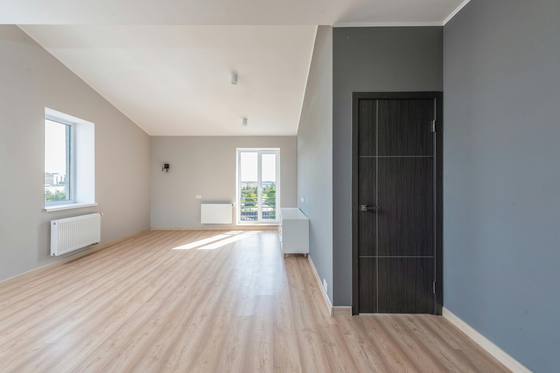 Empty modern room with light wood floors, gray walls, and windows letting in daylight