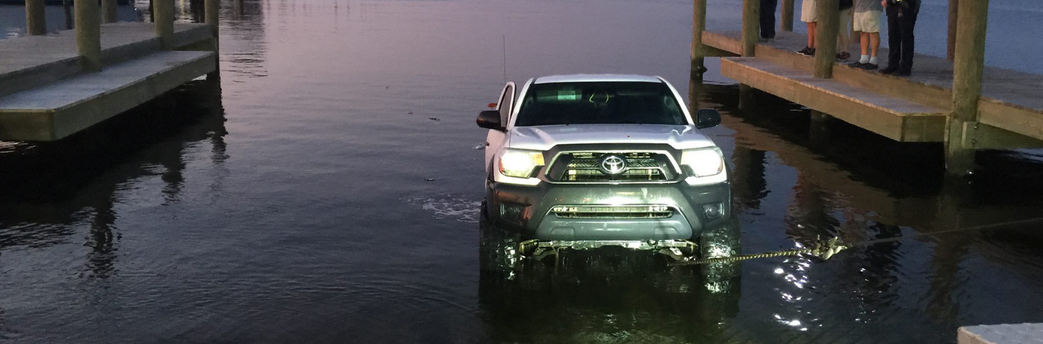 SUV with a chain on the front being pulled up out of water between docked boats at dusk