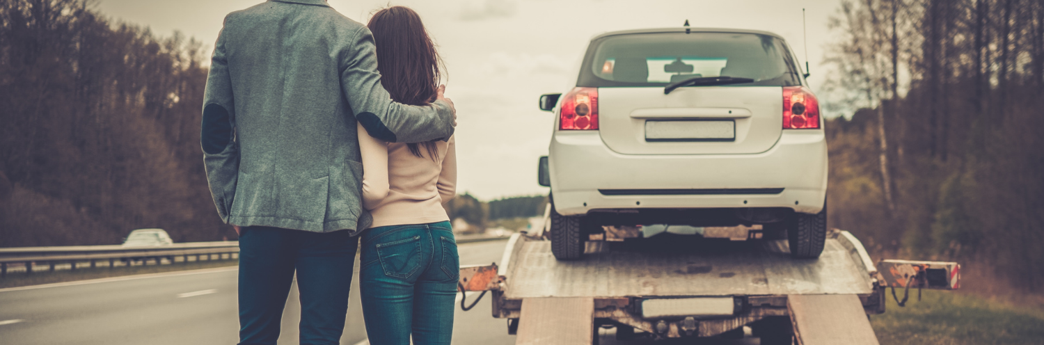 Couple standing by a white car on a roadside, looking on while the car is towed upon a transporting tow trailer