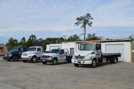 Four white work trucks parked in a lot beside a low warehouse under a cloudy sky