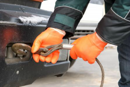 Orange-gloved hands disconnect a tow cable from a car’s front bumper hook on a cloudy day
