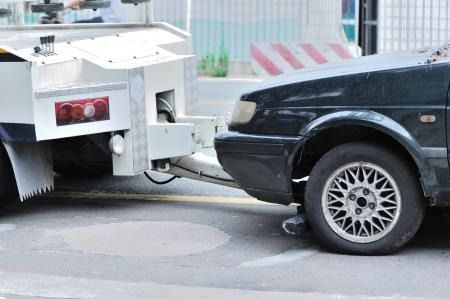 Black car bumper front is attached against the rear of a white tow truck at a street intersection.