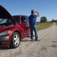Person standing beside a red car with the hood open, and person is looking down the road as if waiting for something on a rural roadside.