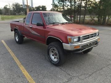 Red pickup truck parked in a lot near trees and a road, viewed from the front-left side