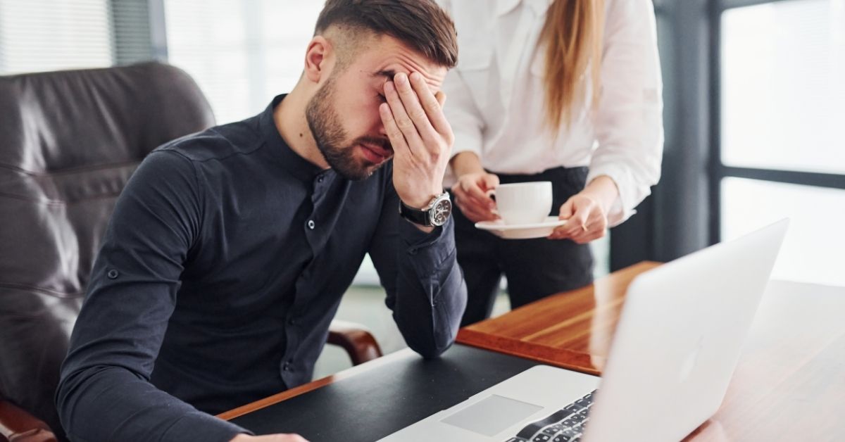 Stressed business owner at his desk with a colleague nearby, representing the frustration of growing