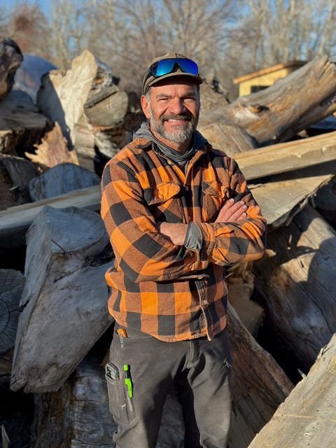 A man in a plaid shirt is standing in front of a pile of wood.
