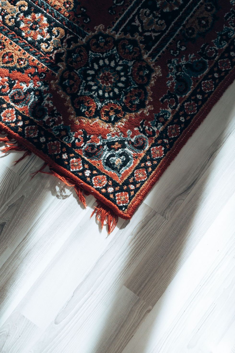 Red patterned rug corner on a light wooden floor with sunlight and fringe detail