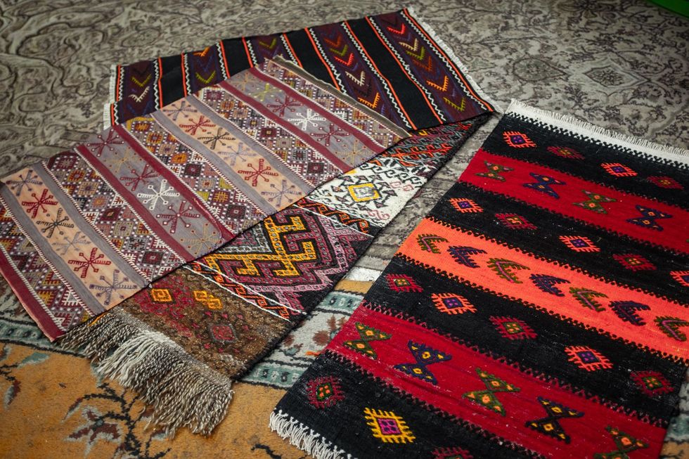 Colorful patterned woven rugs and textiles arranged on a gray stone floor.