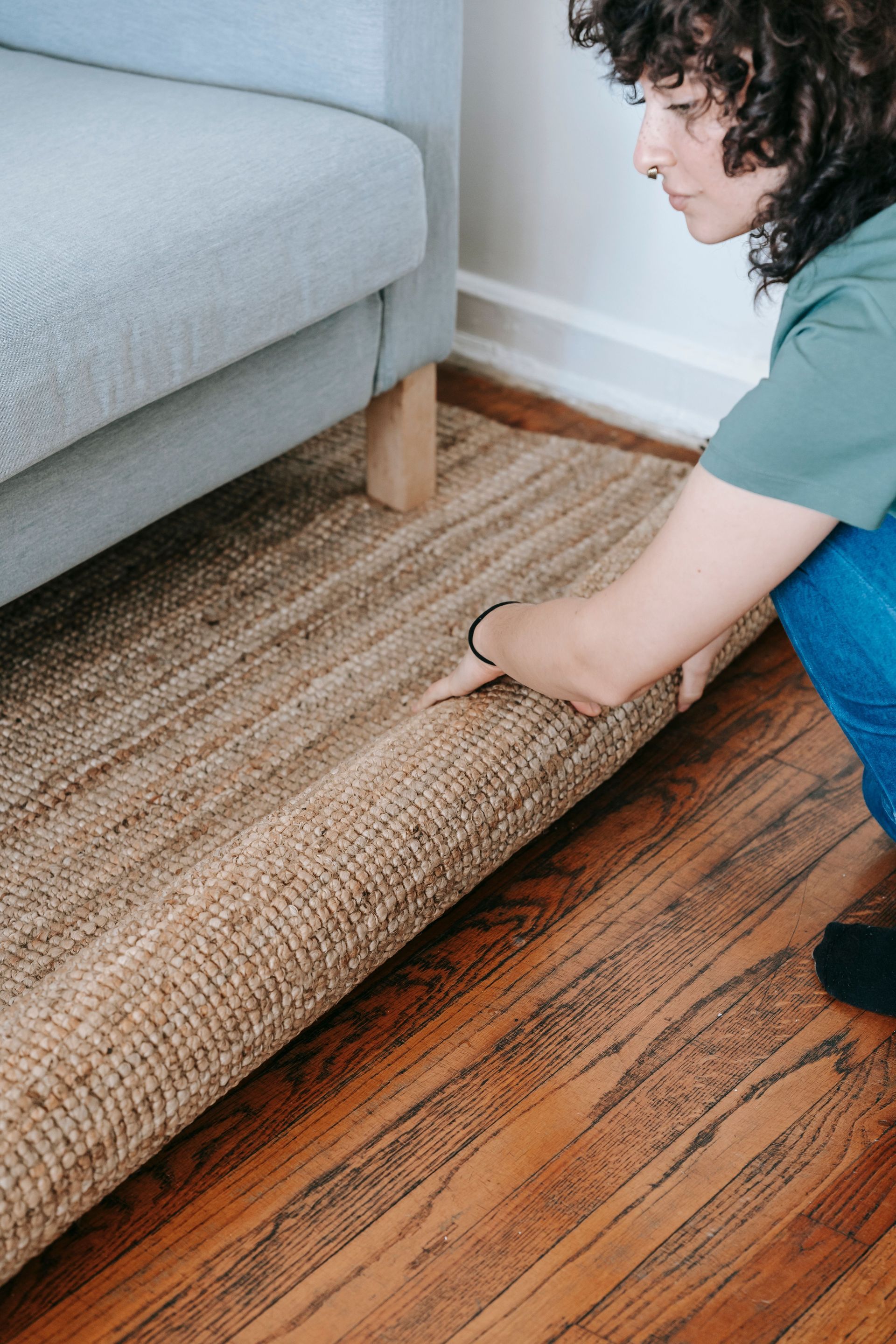 Person rolling up a woven rug beside a gray sofa on a hardwood floor
