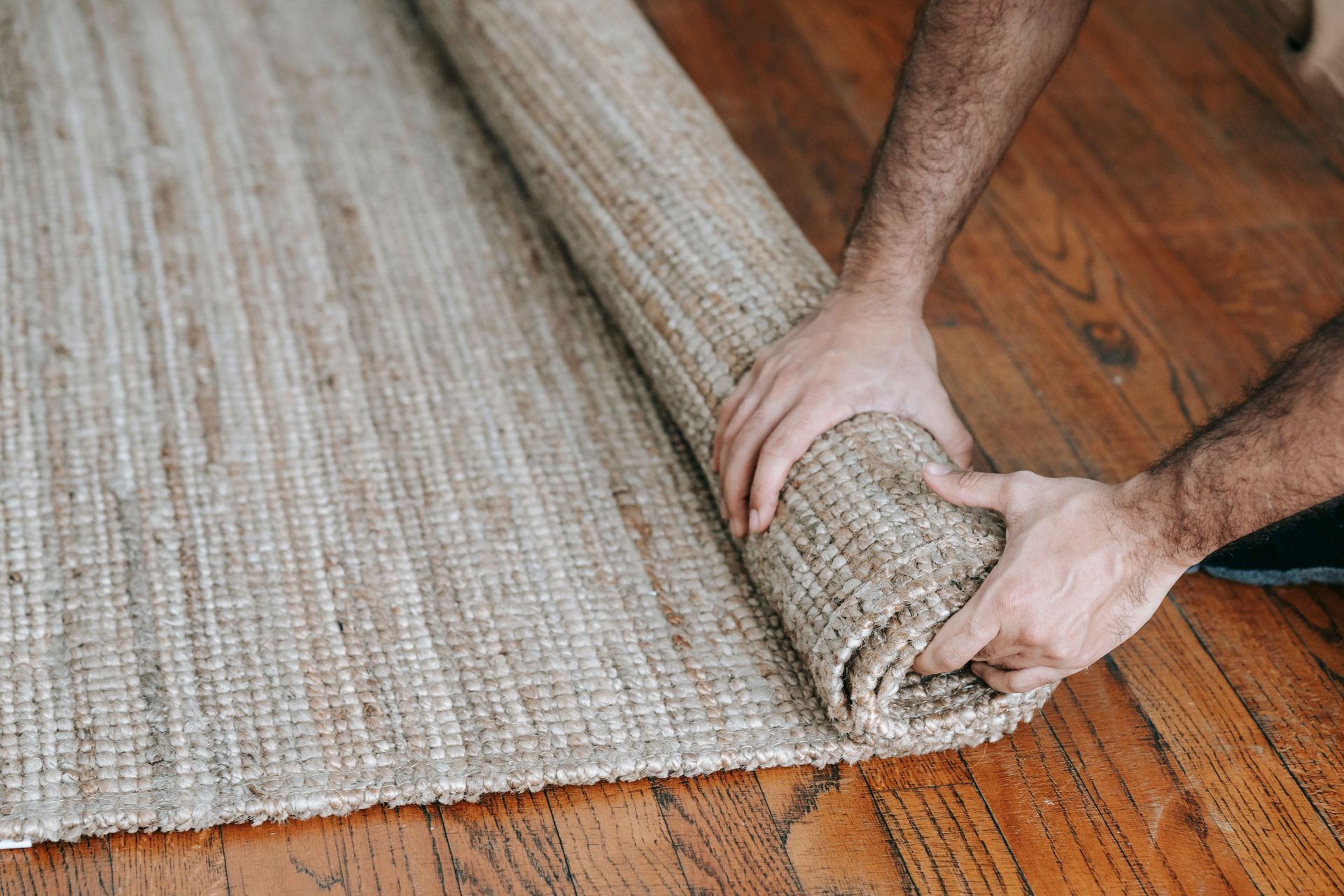 Hands rolling up a beige woven rug on a wooden floor