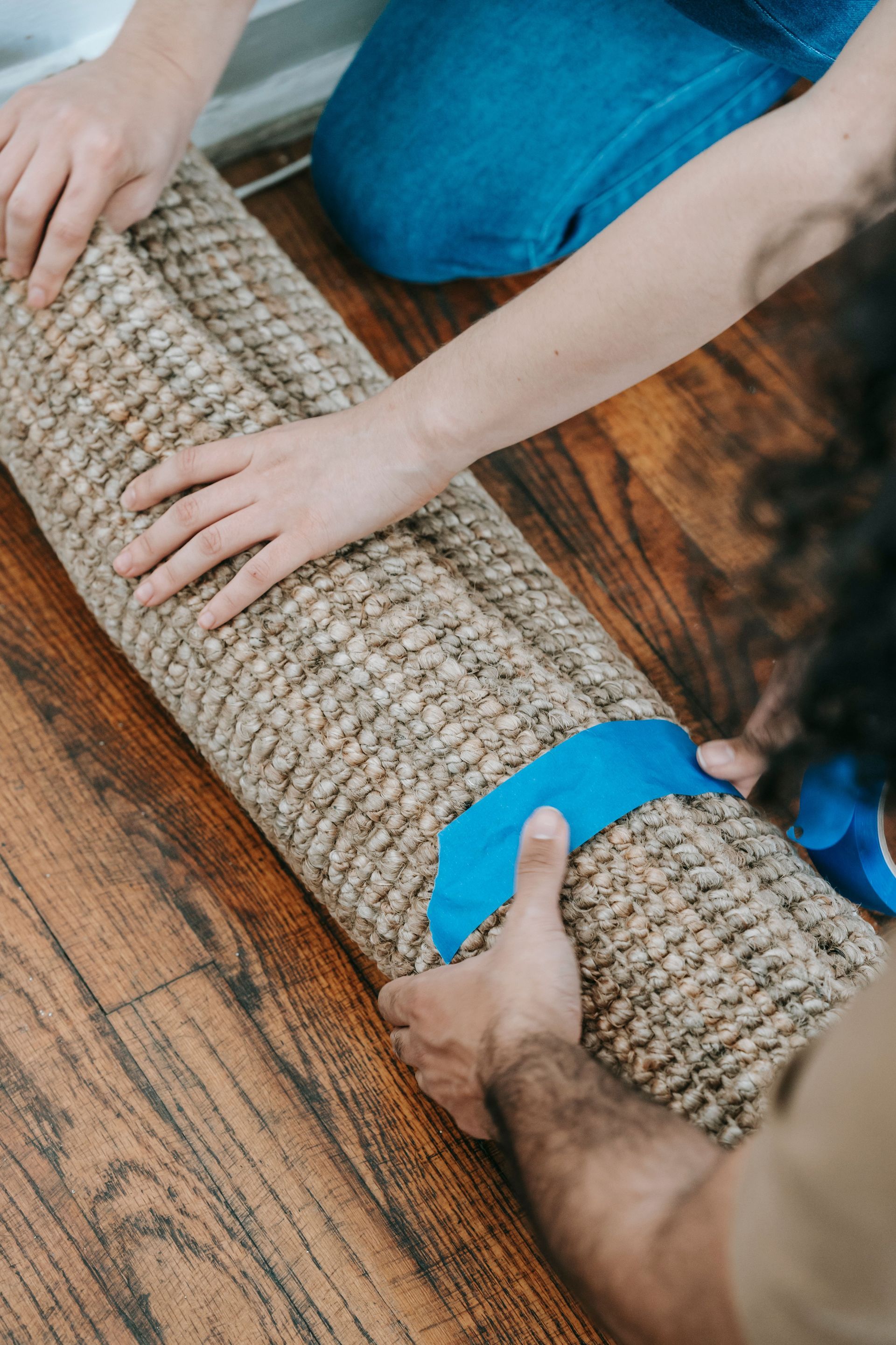 Hands taping a rolled carpet on a wooden floor, with blue painter’s tape.