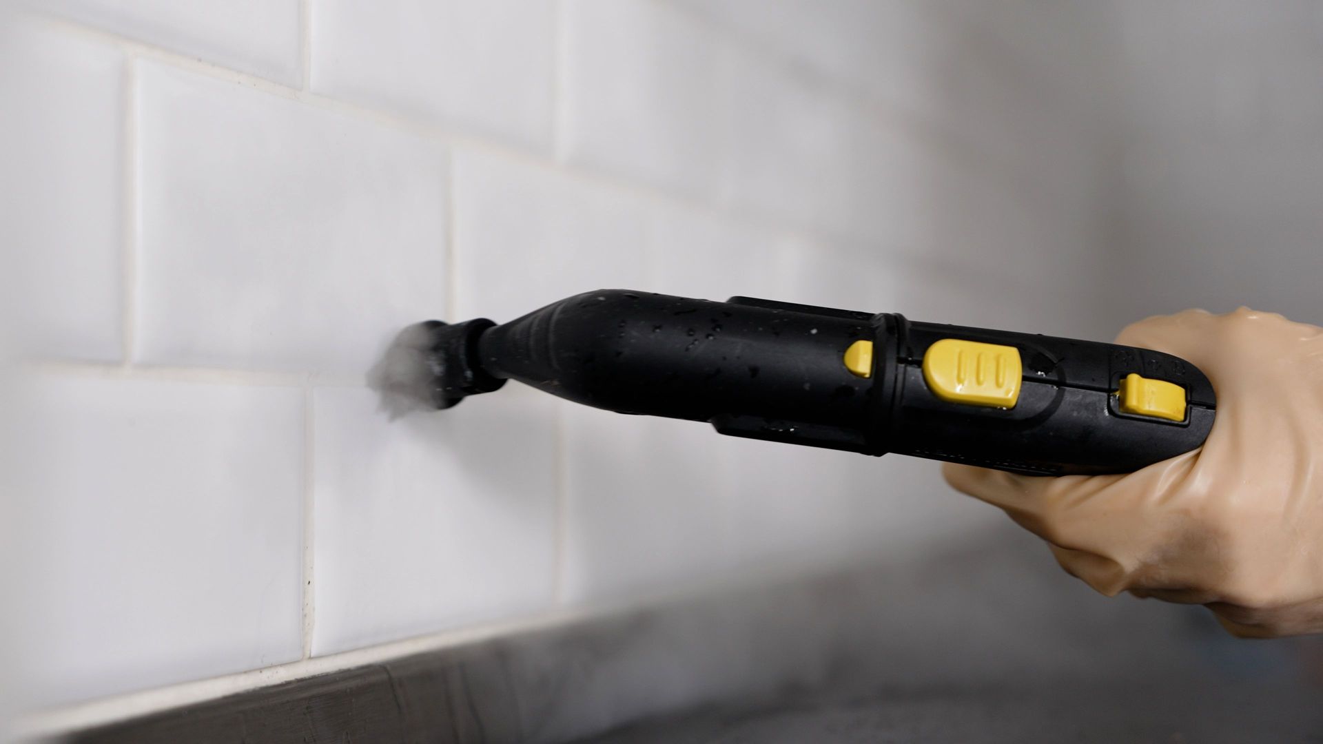 Gloved hand scrubbing a white tiled wall with a green sponge.
