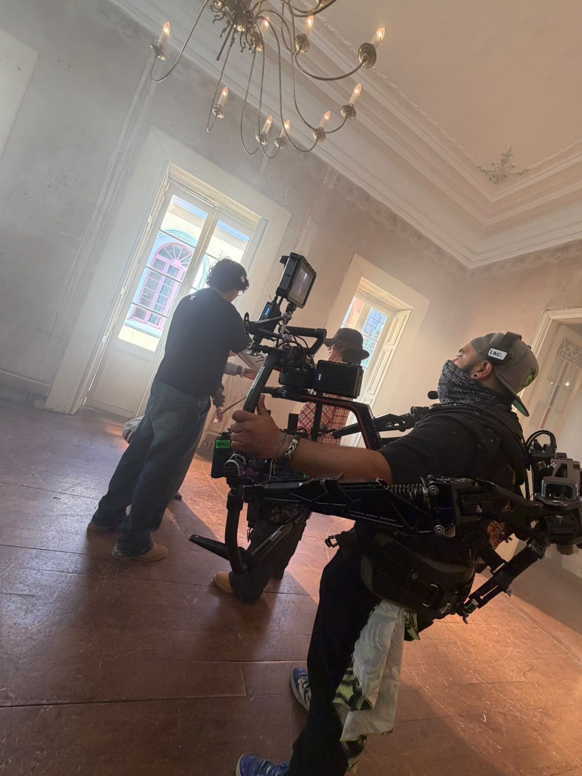 Equipo de cámara filmando en una habitación iluminada por el sol, con ventanas altas y una lámpara de araña