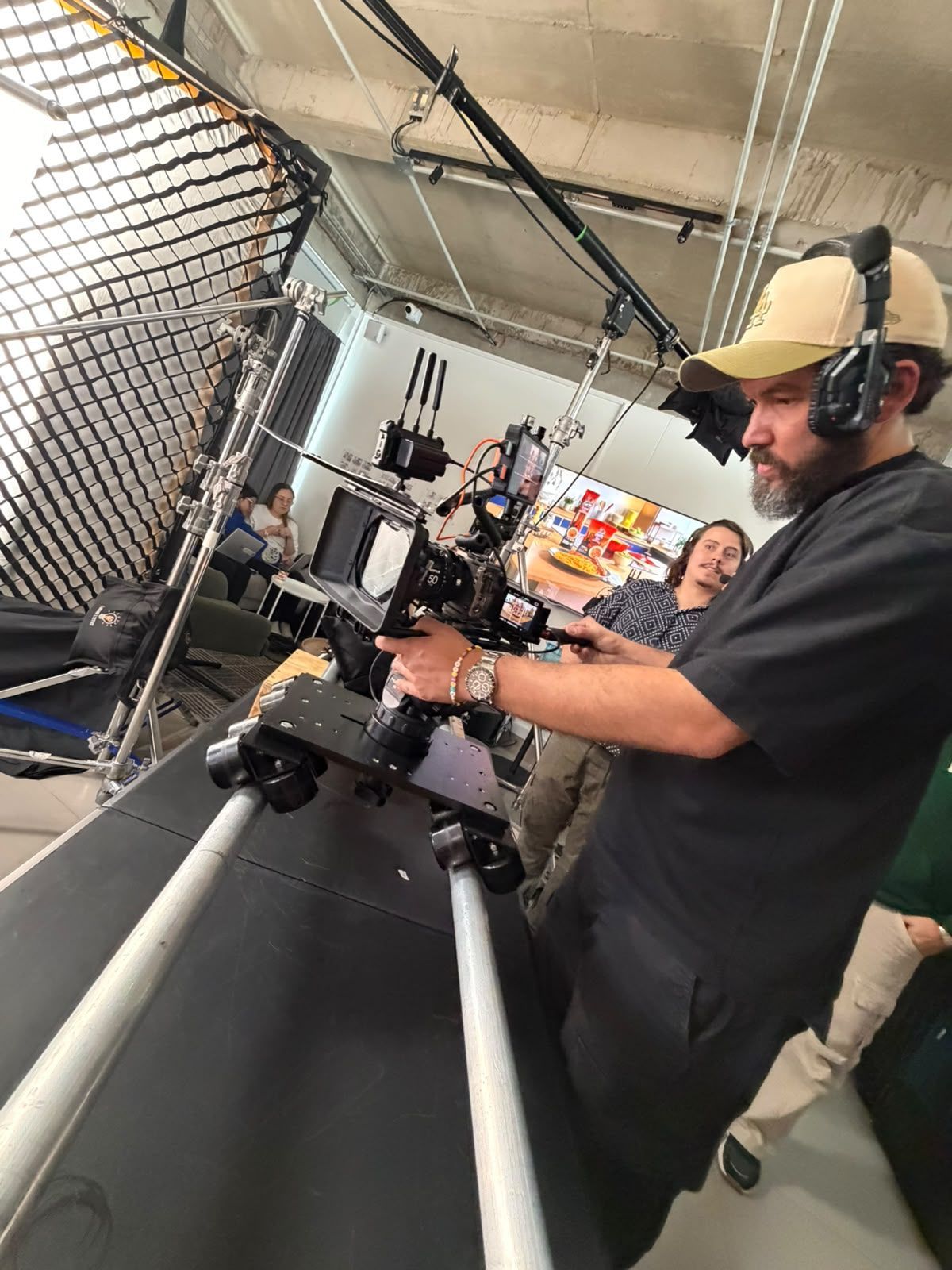 Hombre grabando con un equipo de cámara junto a una valla de malla metálica en un evento en interiores.