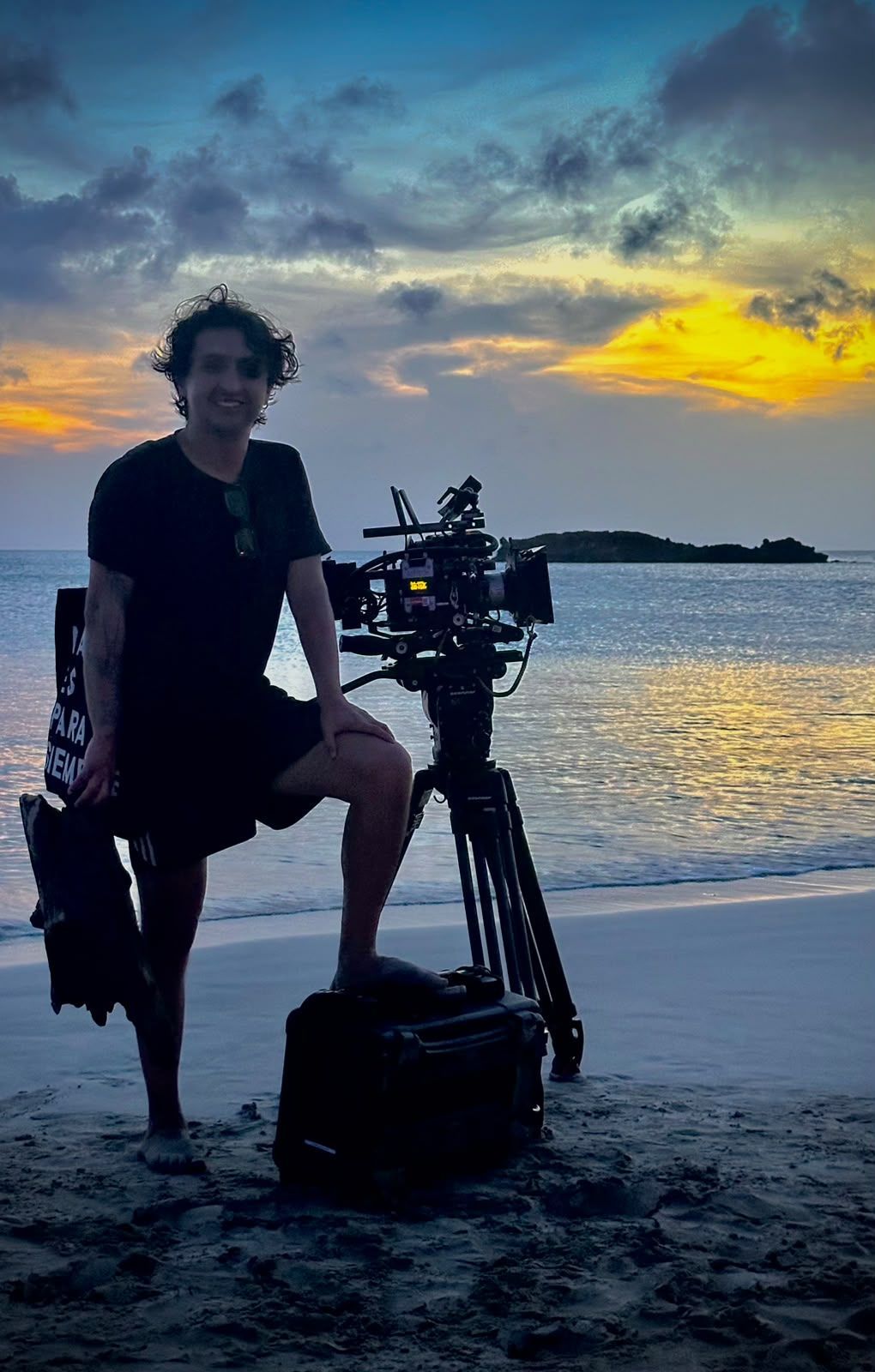 Una persona de pie junto a un equipo de cámara en una playa al atardecer, con nubes coloridas sobre el agua
