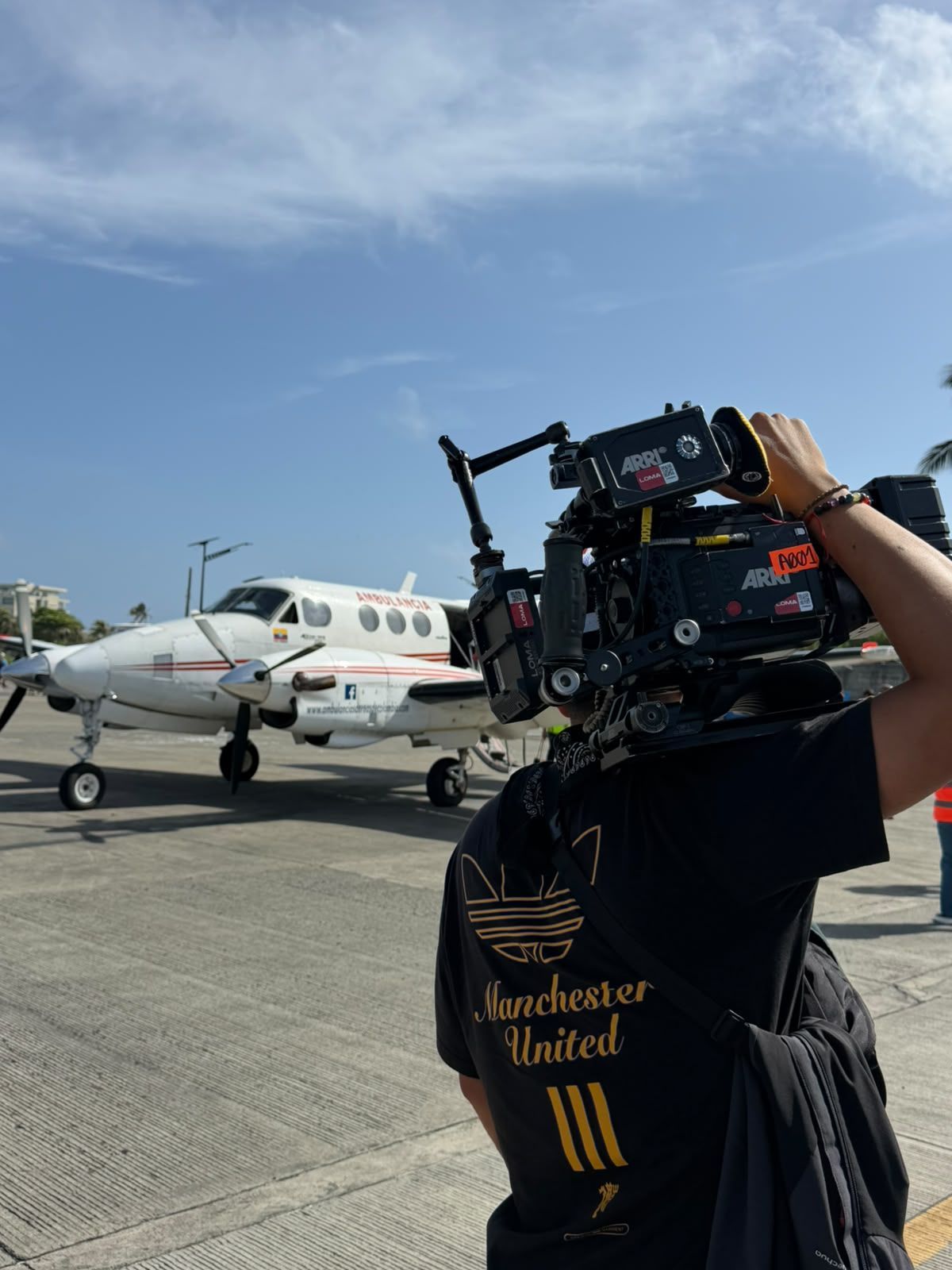 Una persona lleva un equipo de cámara en una pista, con una avioneta y el cielo azul de fondo