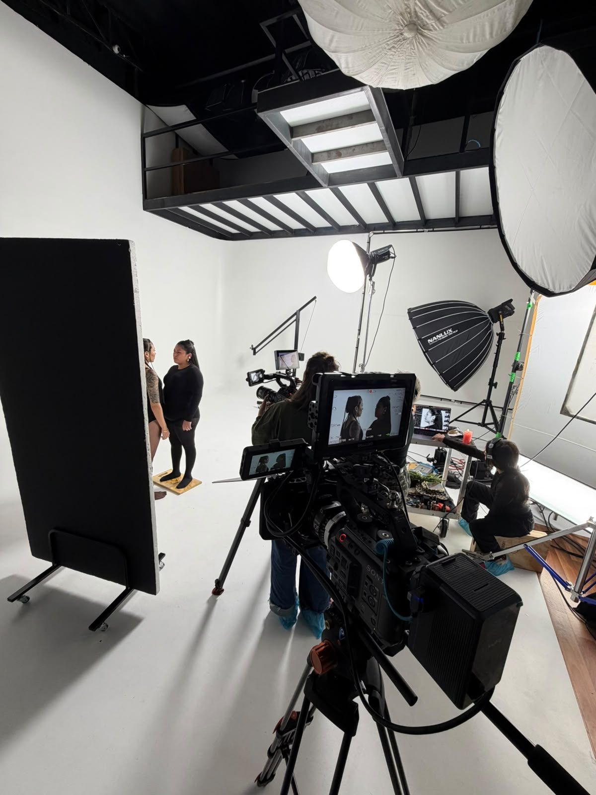 Estudio fotográfico con equipo de cámara, paneles de iluminación y dos personas posando frente a un fondo blanco