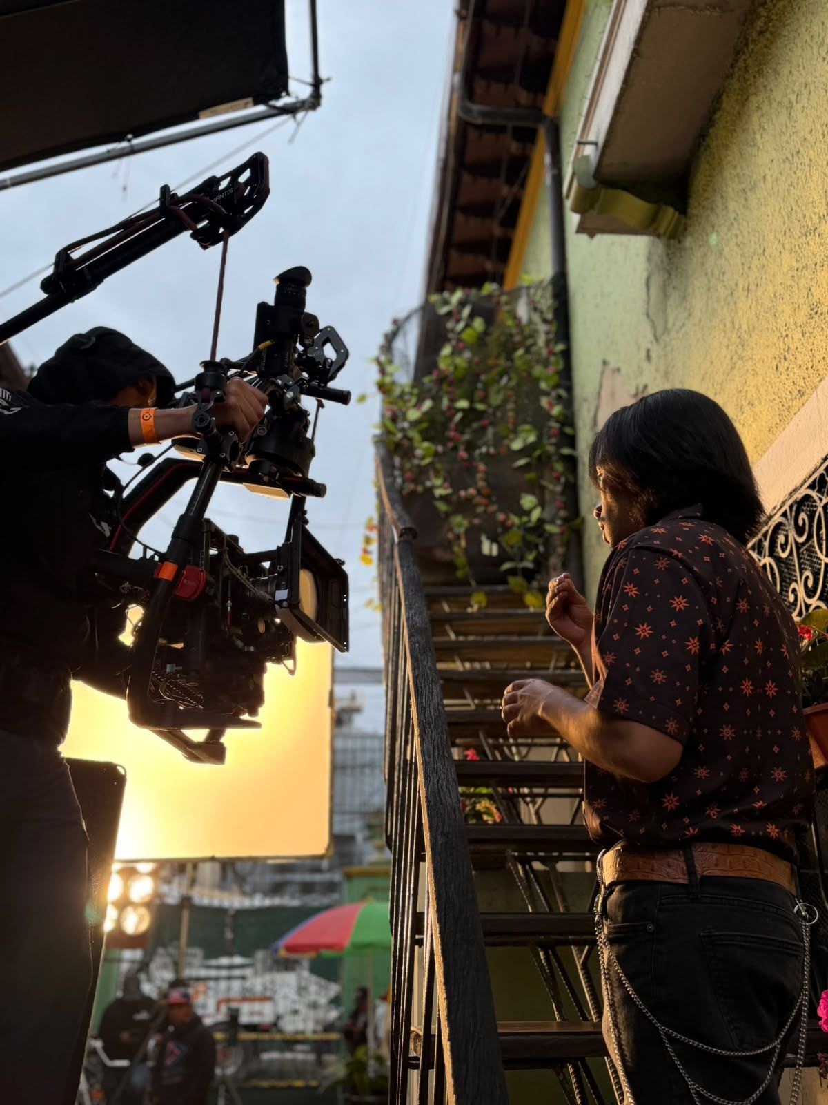 Equipo de rodaje filmando a una mujer en una escalera exterior iluminada por el sol, junto a una pared amarilla