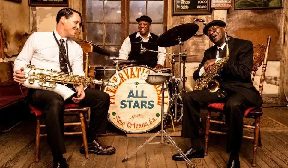 Three men are playing instruments in front of a drum set.