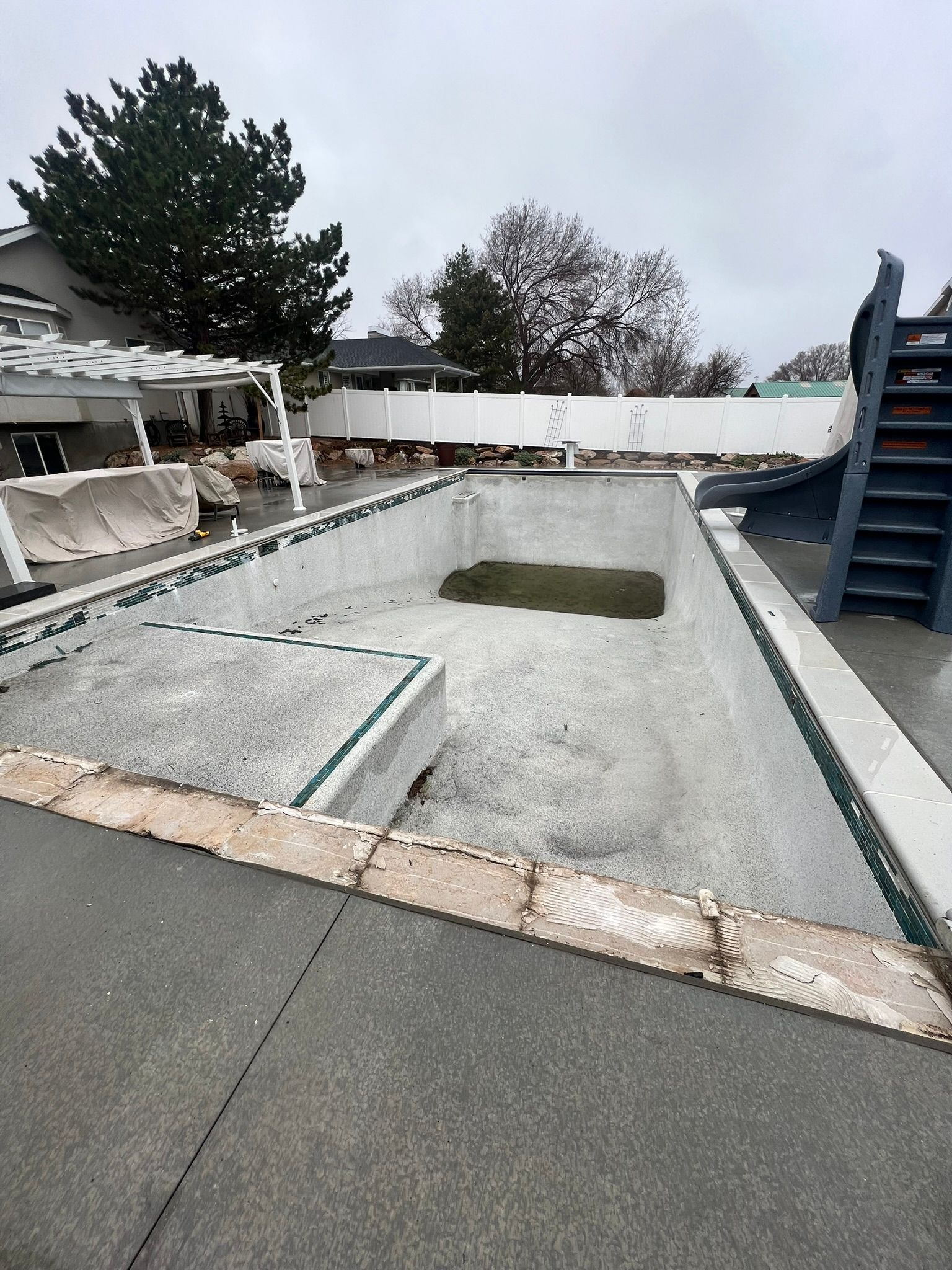 An empty backyard swimming pool under an overcast sky, with a slide on the right and a patio area to the left.