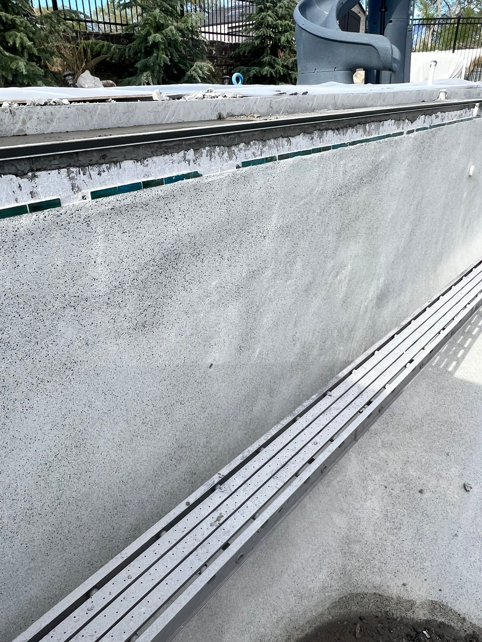 A concrete pool edge under construction, featuring a metal drainage grate and partial decorative tile installation.