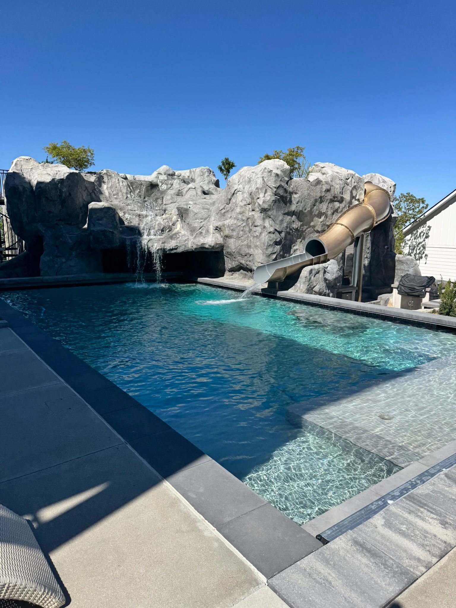 A backyard swimming pool with a large rock formation, a built-in water slide, and a waterfall under a bright blue sky.