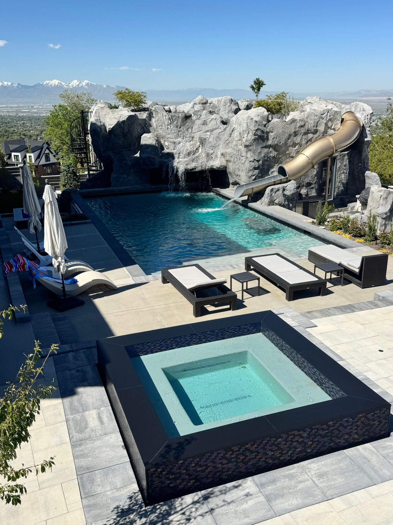 A backyard swimming pool with a rocky waterfall, a slide, a square hot tub, and lounge chairs under a clear blue sky.
