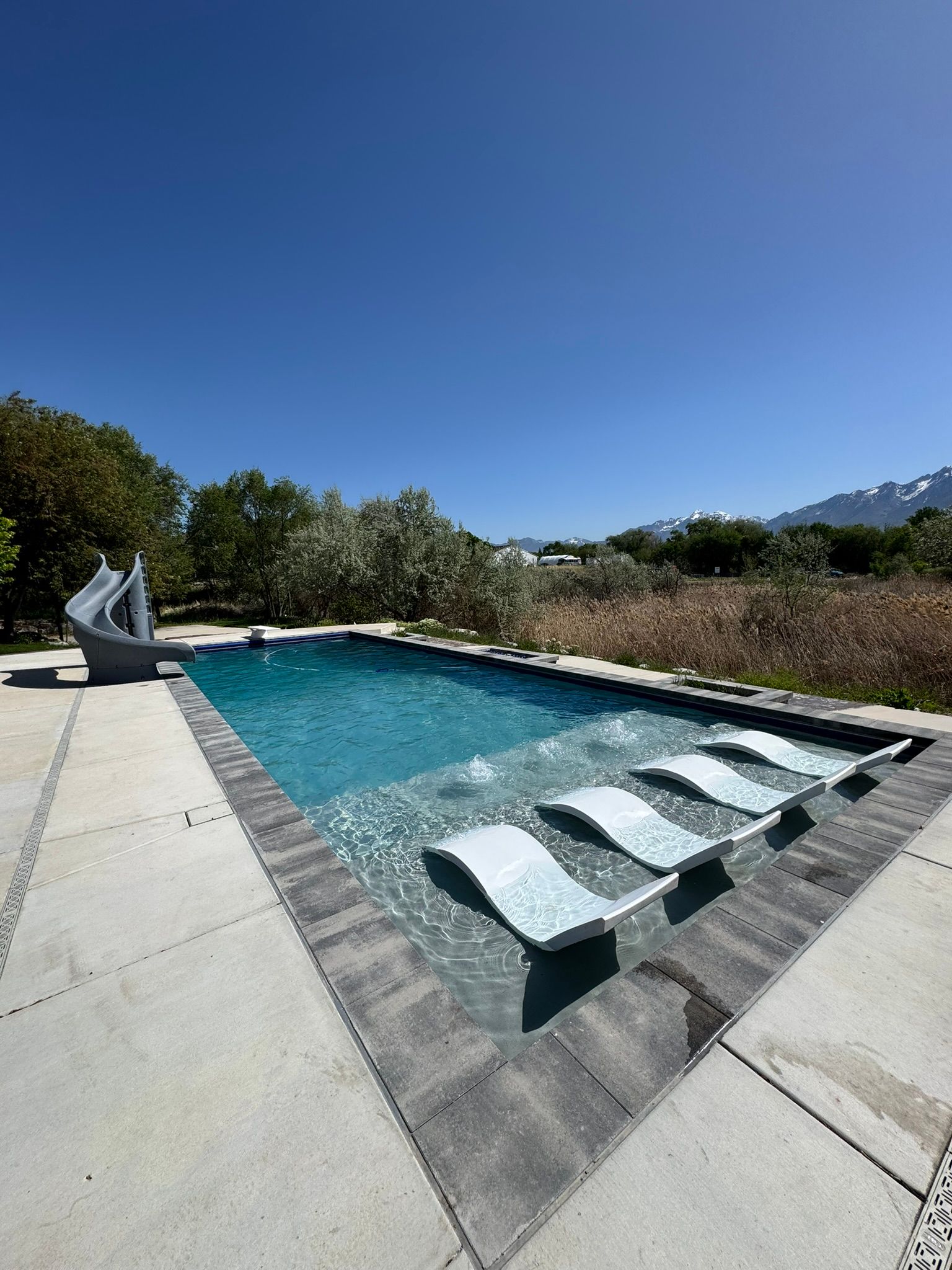 A swimming pool featuring a shallow tanning ledge with three lounge chairs, a slide, and a mountain backdrop.