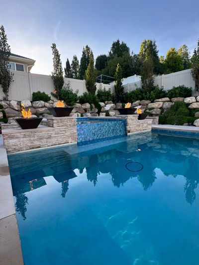 A blue backyard swimming pool featuring a waterfall wall with three lit fire bowls on a stone ledge under a clear sky.