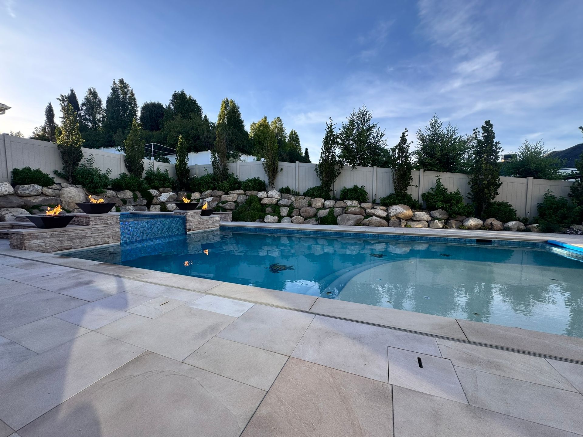 A luxury backyard pool with stone decking, a blue tiled water feature, and burning fire bowls set against a stone wall.