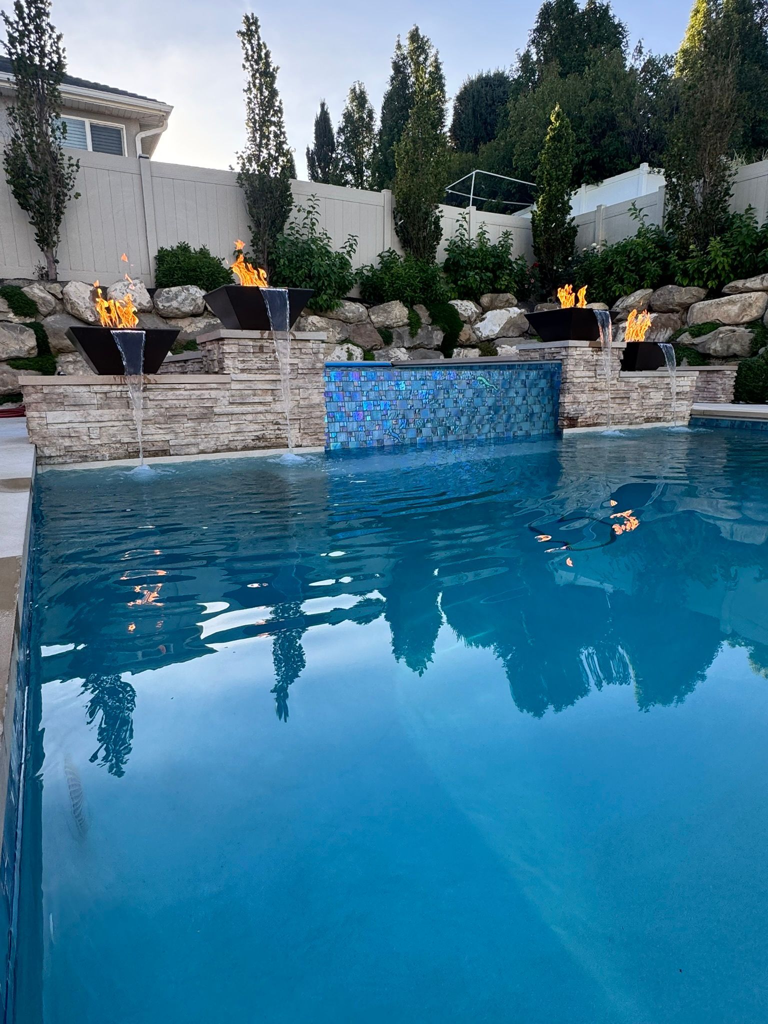 A blue swimming pool with a stone wall feature, decorative blue tiles, and four glowing fire bowls at dusk.