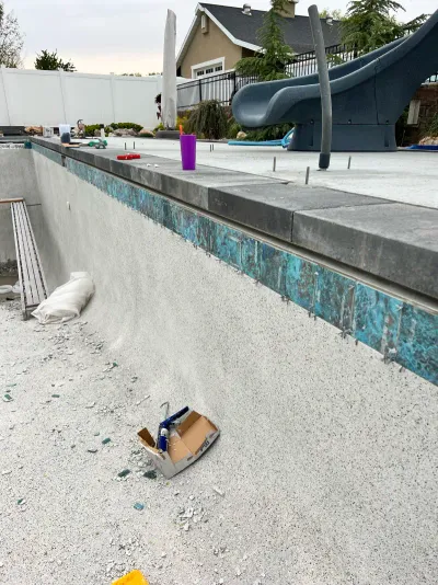 An empty swimming pool under construction, featuring gray stone coping, blue patterned tile, and a slide in the background.