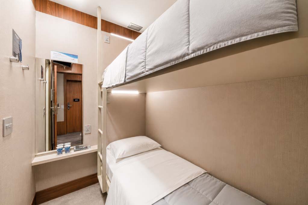 Quarto de hotel compacto com beliches brancos, pia pequena, espelho e parede e porta com painéis de madeira.
