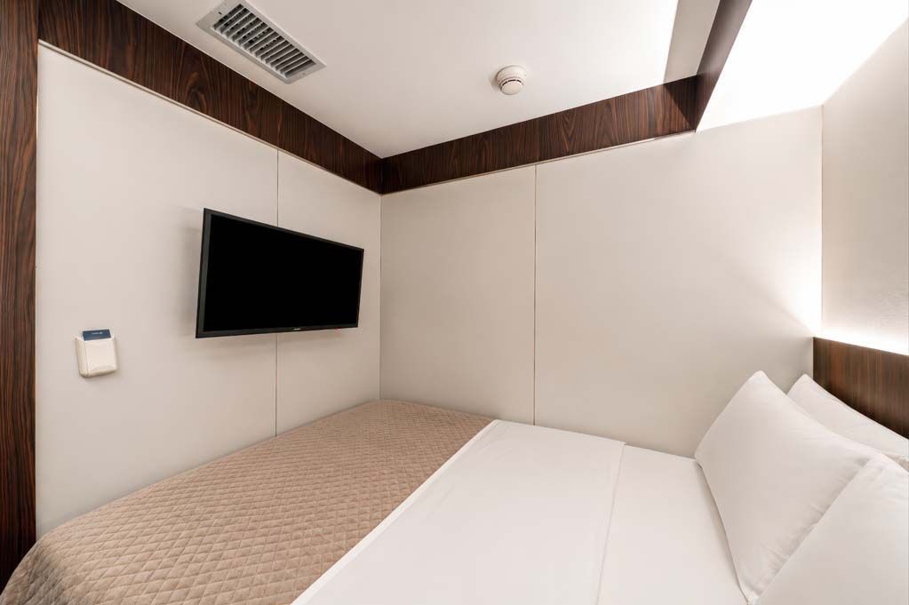 Quarto de hotel pequeno com duas camas de solteiro, TV de parede e paredes acolchoadas bege.