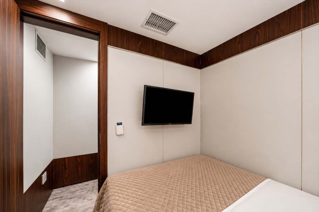 Quarto pequeno com cama, TV de parede e porta aberta para o banheiro ou corredor.