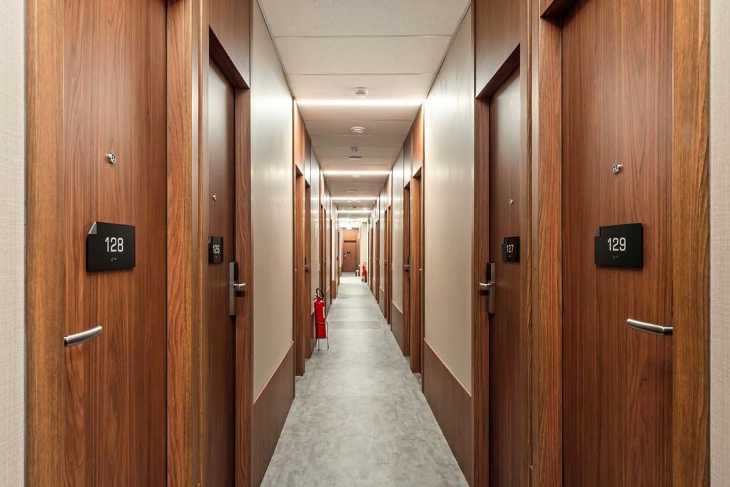 Longo corredor com portas de apartamentos em painéis de madeira e carpete bege, visto da linha central.