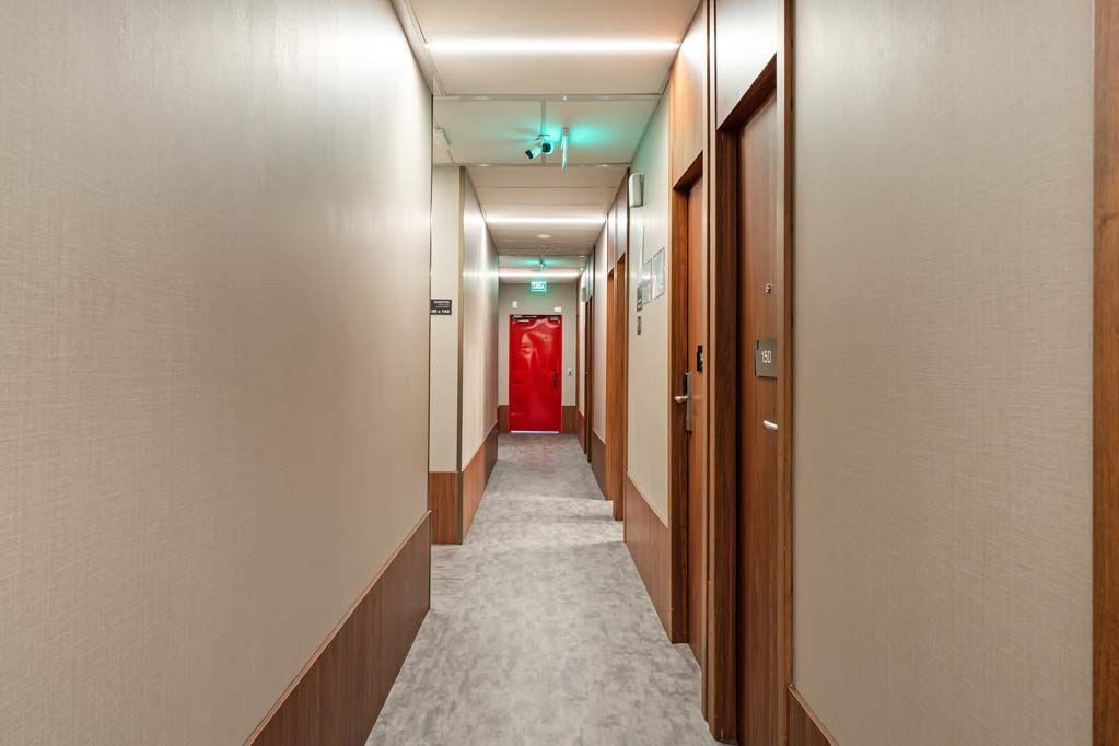 Longo corredor de hotel com paredes bege, portas de madeira, carpete cinza e uma porta de saída vermelha no final.