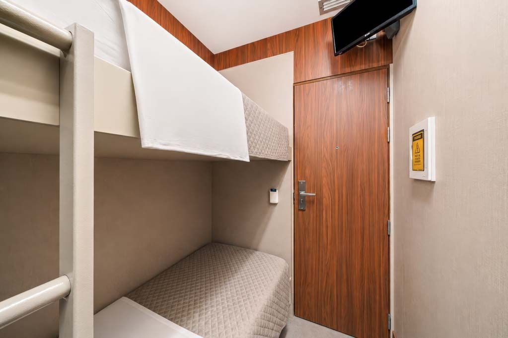 Cabine compacta com beliche, porta de madeira, paredes bege e uma pequena TV instalada perto do teto.
