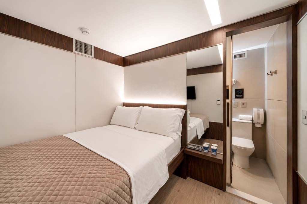 Quarto de hotel compacto com cama, espelho e banheiro com box de vidro.