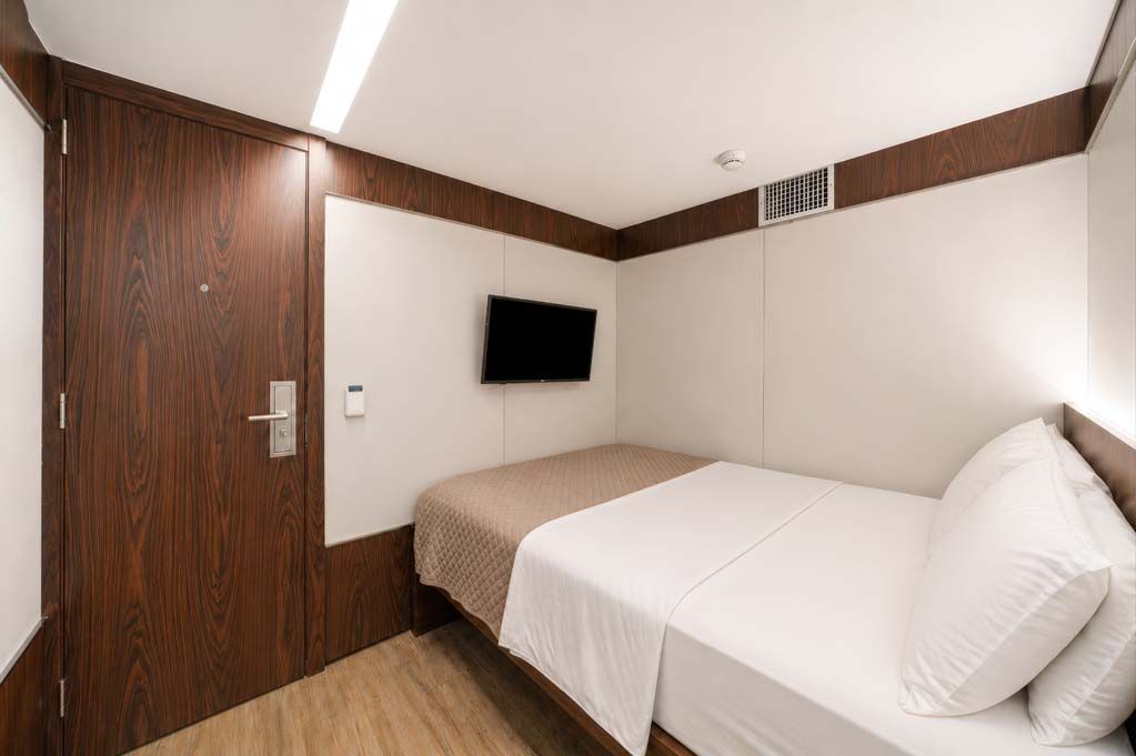 Quarto de hotel pequeno com uma cama de solteiro, TV de parede, porta de madeira e paredes brancas.