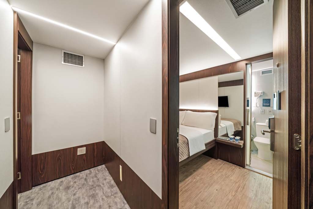 Corredor estreito e quarto pequeno com paredes bege, acabamento em madeira, cama e banheiro visíveis através de uma porta aberta.