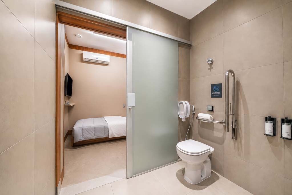 Banheiro acessível no quarto de hotel com vaso sanitário, barras de apoio, pia e porta de correr fosca para a área da cama.