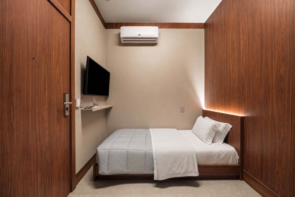 Quarto pequeno e moderno com cama de solteiro, TV de parede, paredes com painéis de madeira e ar-condicionado.