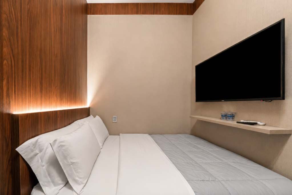 Quarto de hotel moderno com cama, parede revestida de madeira e TV fixada na parede em uma pequena prateleira.