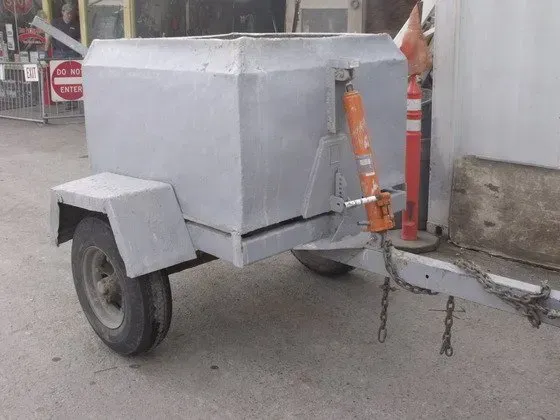 Gray metal trailer with box-shaped compartment and wheels parked outside a building.