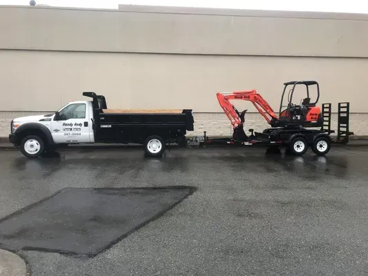 White truck towing a trailer with a small orange excavator parked on wet asphalt.