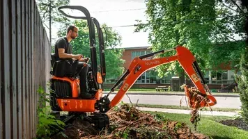 Man operating a Kubota mini-excavator, digging near a wooden fence and trees, outdoors.